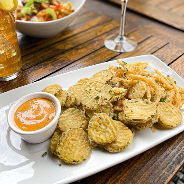 Crispy fried pickles and onion straws with a side of dipping sauce on a white plate.