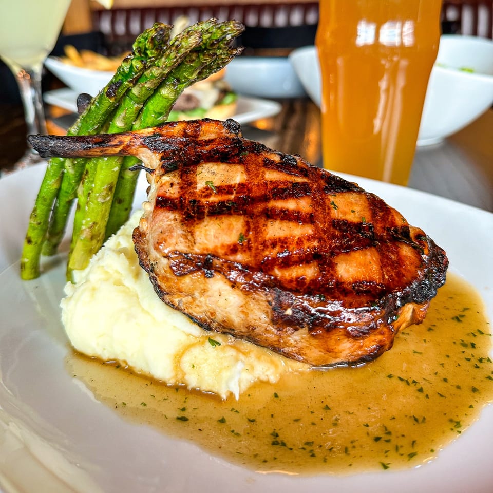 Grilled pork chop with mashed potatoes, asparagus, and gravy, with drinks in the background.