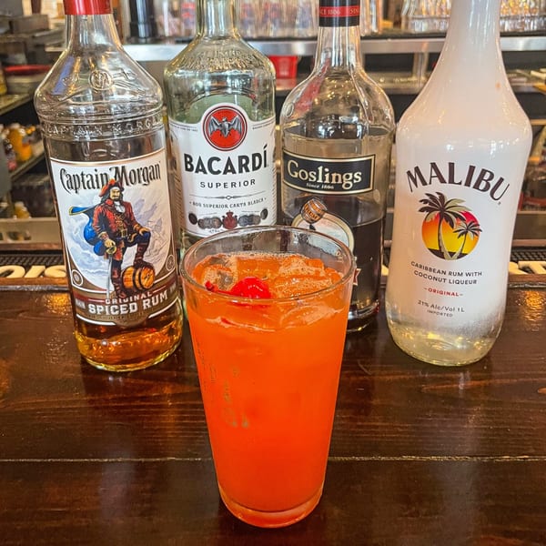 A vibrant orange cocktail with a cherry garnish, surrounded by bottles of rum on a wooden bar.