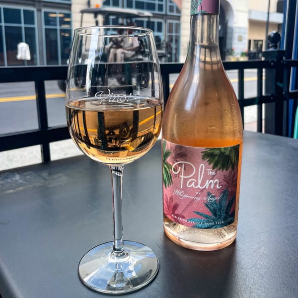 A glass of rosé wine and a bottle of The Palm by Whispering Angel on an outdoor table.