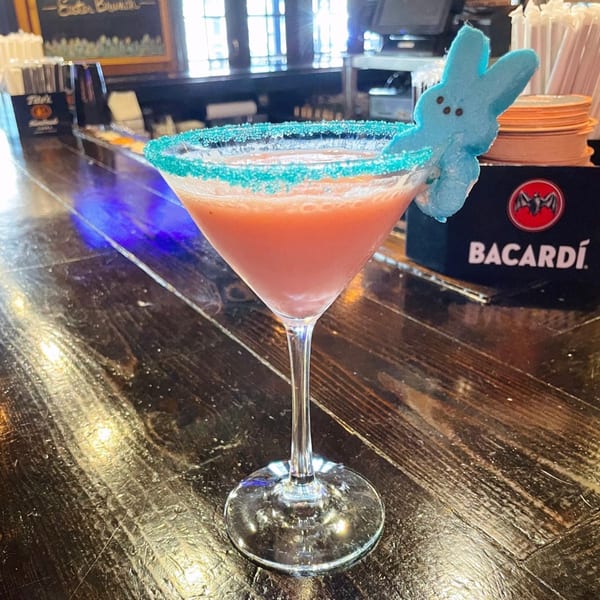 A pink cocktail with a blue sugar rim and a blue Peeps bunny garnish on a wooden bar.