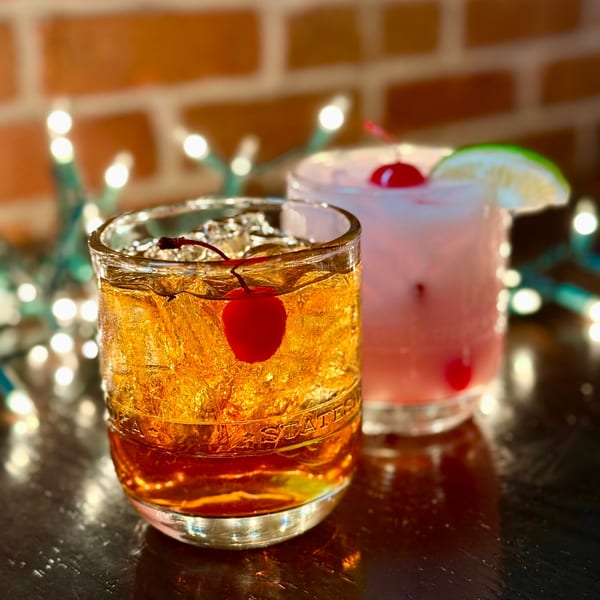 Two cocktails, one amber with a cherry and one pink with a lime, on a bar.