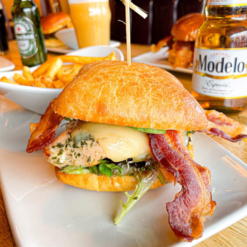 A chicken sandwich with melted cheese, bacon, and lettuce, served with fries and beer.