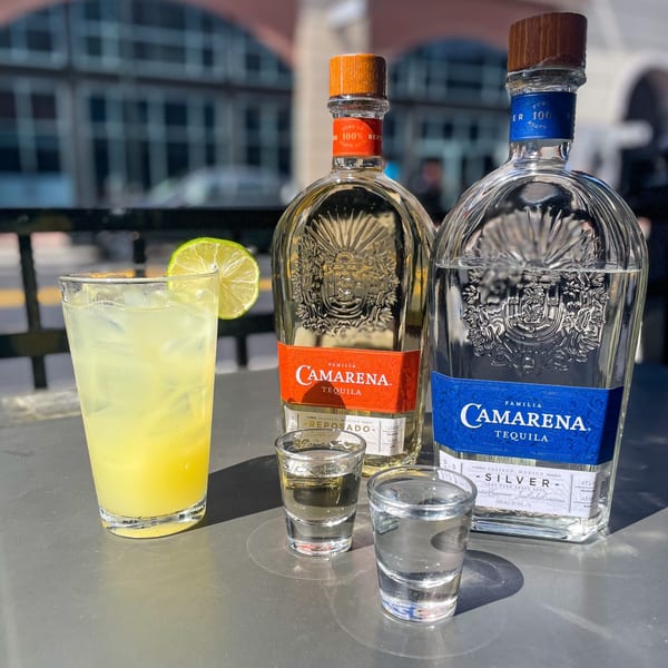 Two bottles of Camarena Tequila, a cocktail with lime, and two shot glasses on an outdoor table.