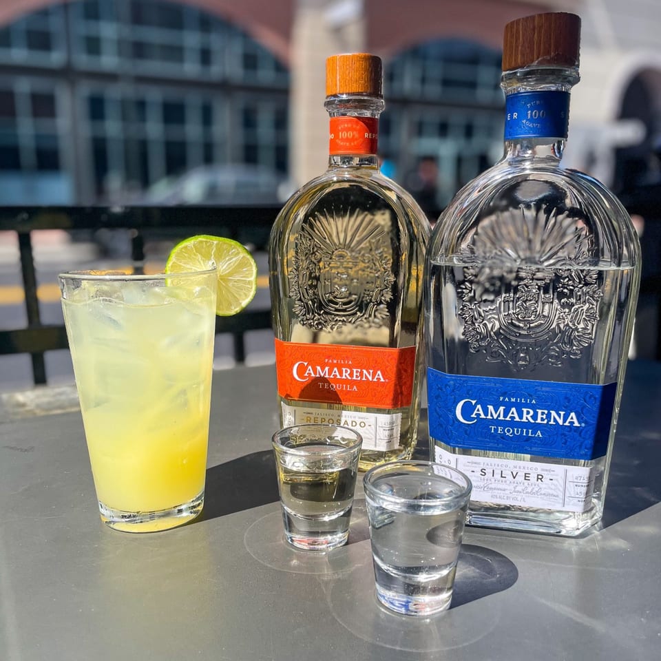 Two bottles of Camarena Tequila, a cocktail with lime, and two shot glasses on an outdoor table.