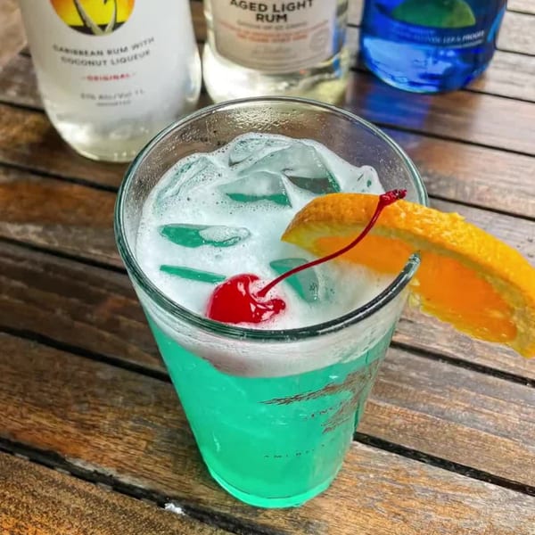 A blue cocktail with foam, ice, an orange slice, and a cherry, with liquor bottles in the background.